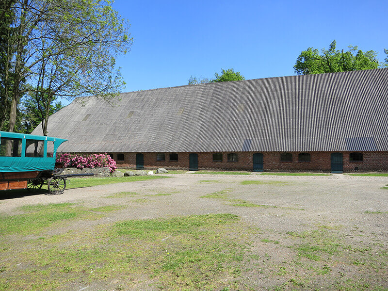 Das Kuhhaus von der Seite mit dem Vorplatz und einem Pferdeanhänger
